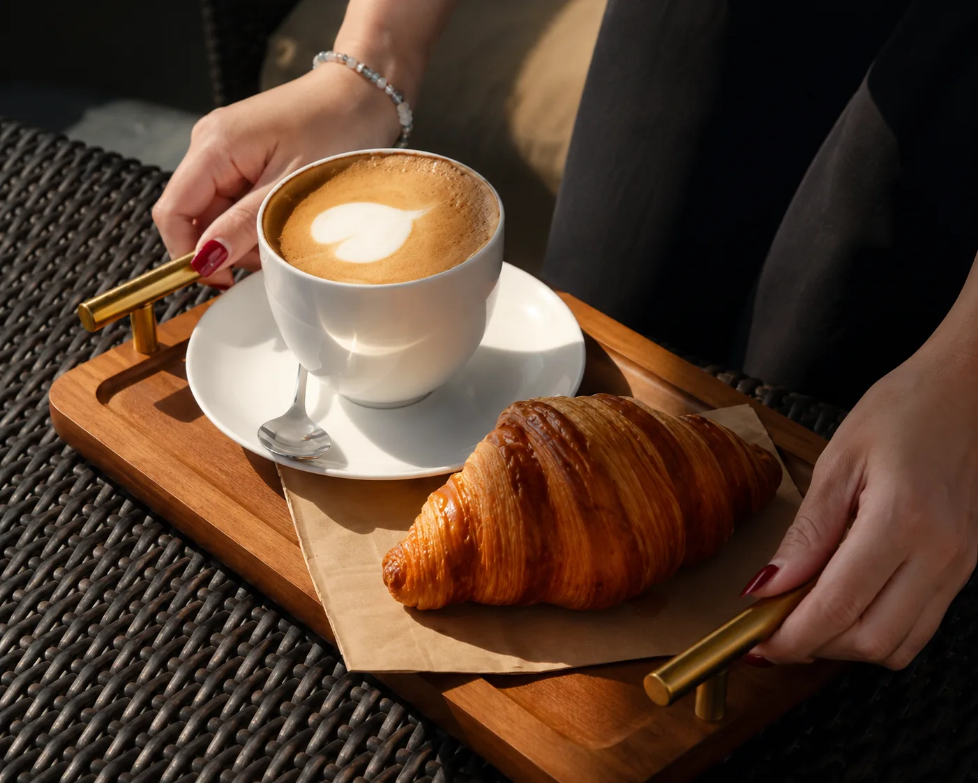 Hands holding a wooden tray with a latte and a fresh croissant in warm sunlight